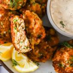 Plate of crab cakes garnished with lemon wedges and dill, served with a bowl of dipping sauce. Text on image reads “Old Bay Crab Cakes” and “LenasKitchenBlog.com.”.