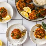 Four plates with crab cakes, garnished with fresh herbs and lemon wedges, are arranged on a white tiled surface with a bowl of dipping sauce nearby.