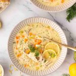 A bowl of Greek lemon chicken soup garnished with lemon slices and dill, surrounded by chopped vegetables, bread, and fresh herbs on a white surface.