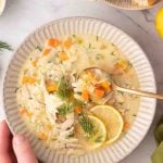 A bowl of avgolemono Greek lemon chicken soup with shredded chicken, carrots, orzo, dill, and lemon slices, served with bread on the side.