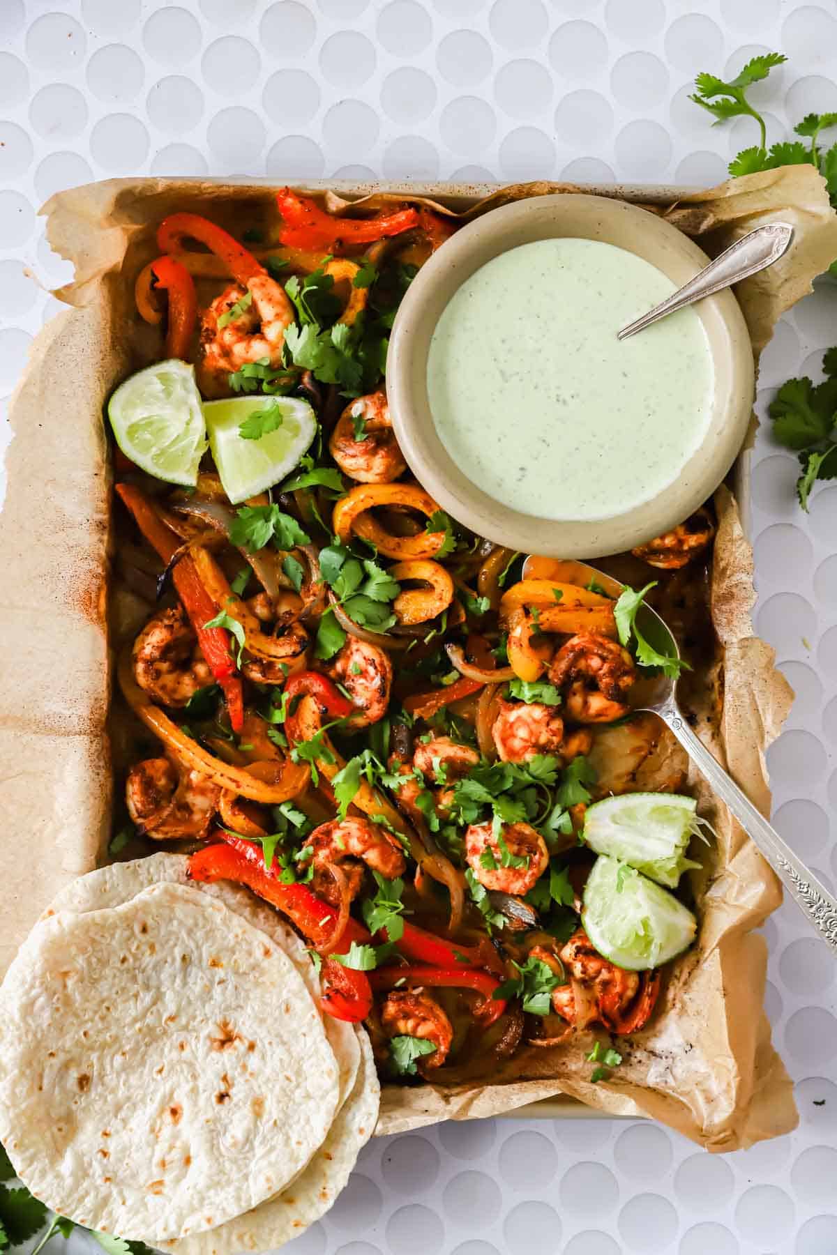 A tray of shrimp fajitas with bell peppers, onions, lime wedges, fresh cilantro, flour tortillas, and a bowl of green sauce on the side.