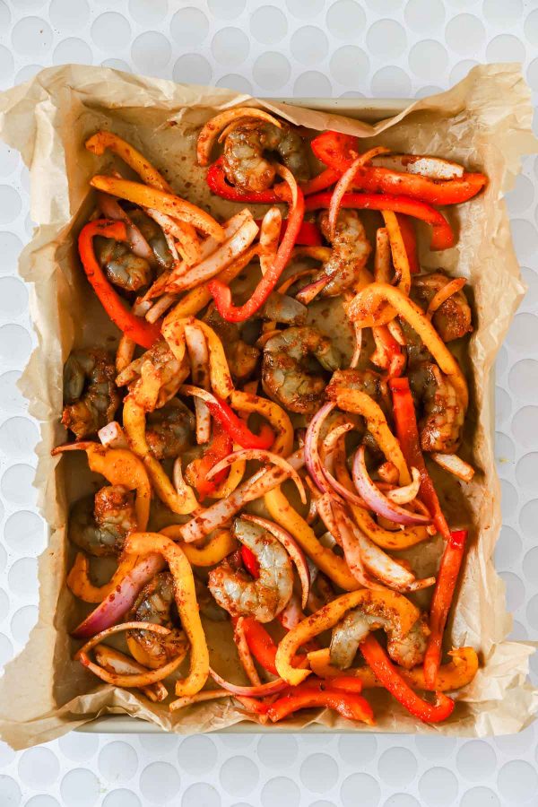 Raw shrimp, sliced red and yellow bell peppers, and red onion arranged on a parchment-lined baking sheet, seasoned with spices, ready to be cooked.