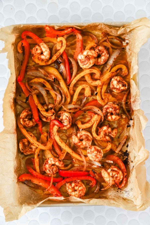 Baking tray lined with parchment paper containing cooked shrimp, sliced red bell peppers, and onions seasoned with spices.