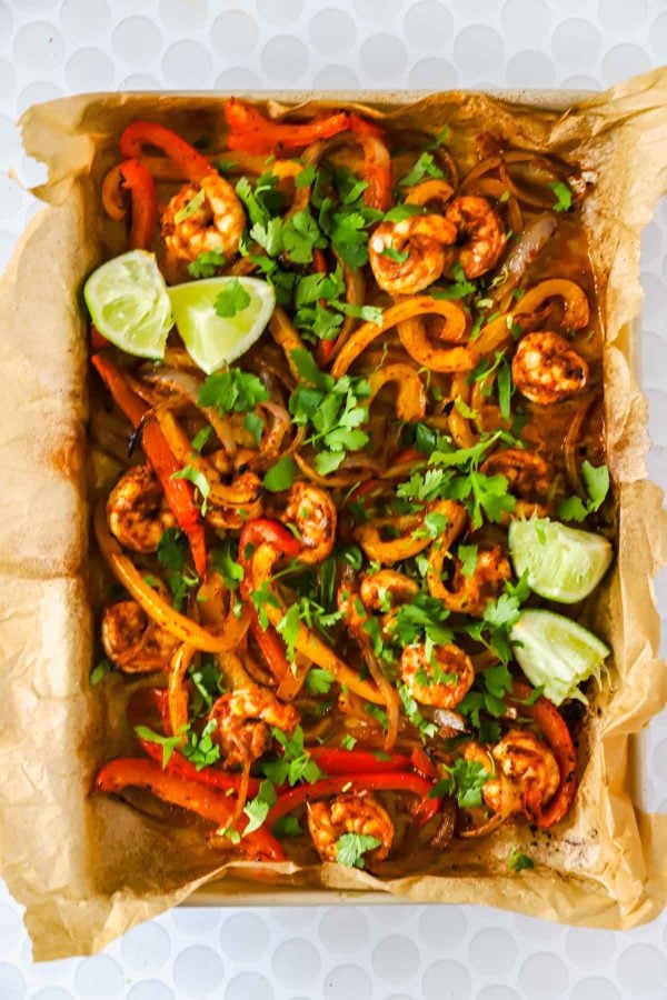 Sheet pan with baked shrimp, sliced bell peppers, onions, fresh cilantro, and lime wedges on parchment paper.
