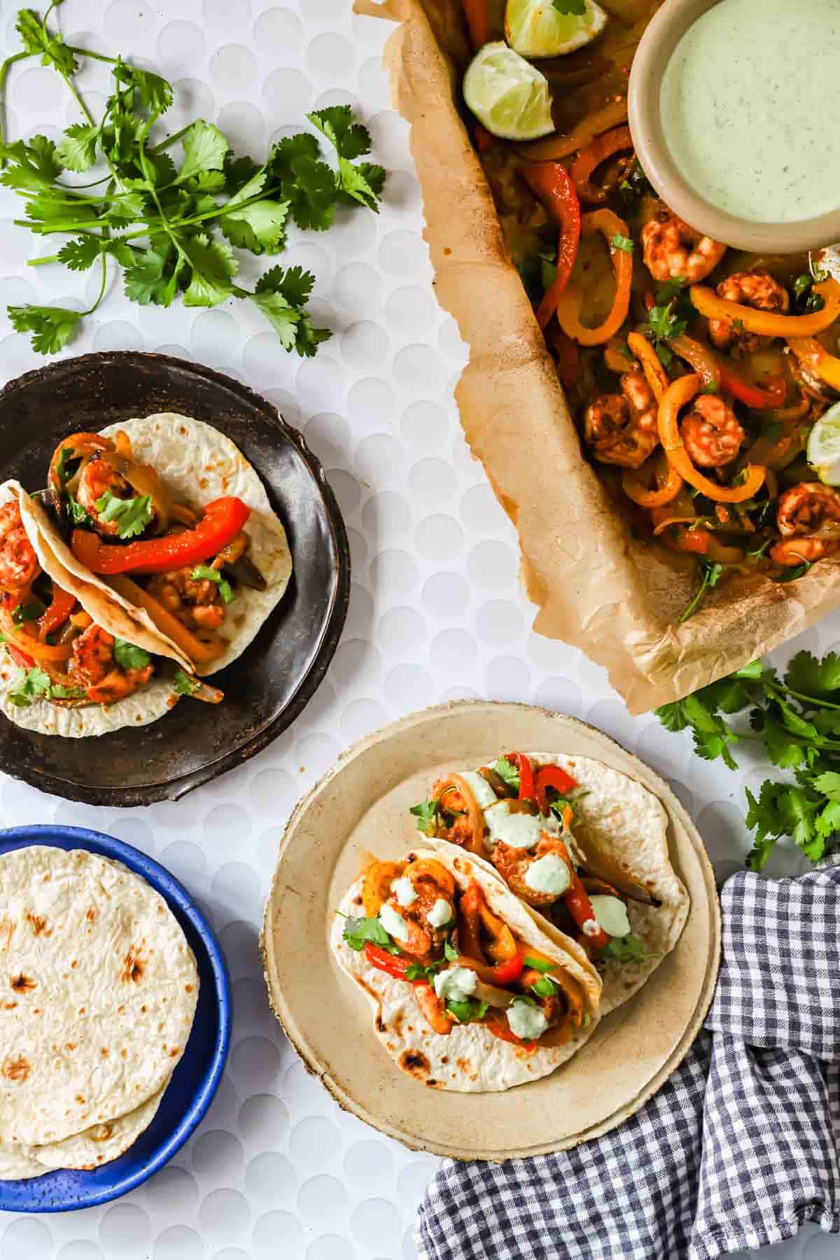 Three plates with tortillas and vegetable tacos, a baking dish of taco filling with lime wedges, fresh cilantro, and a checkered cloth on a white surface.
