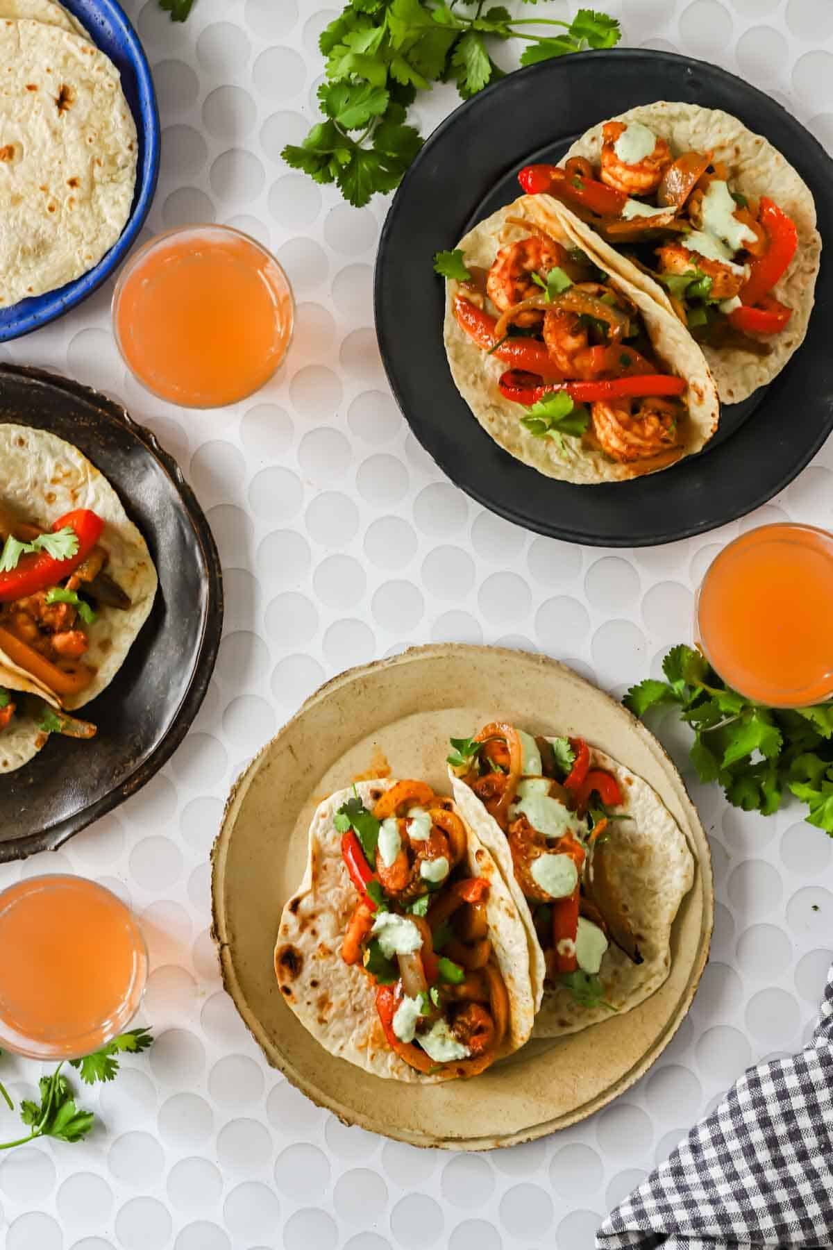 Three plates of shrimp fajita tacos with bell peppers and sauce, surrounded by three glasses of pink drink, cilantro, and tortillas on a white textured surface.