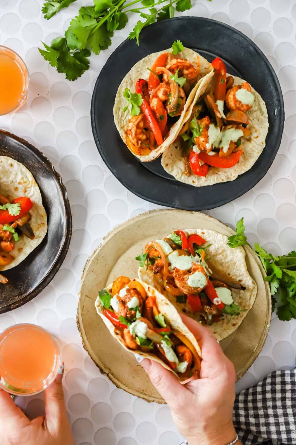 Three plates with soft tacos filled with grilled shrimp, red bell peppers, herbs, and a creamy sauce. A hand holds one taco and two glasses of pink beverage are nearby.