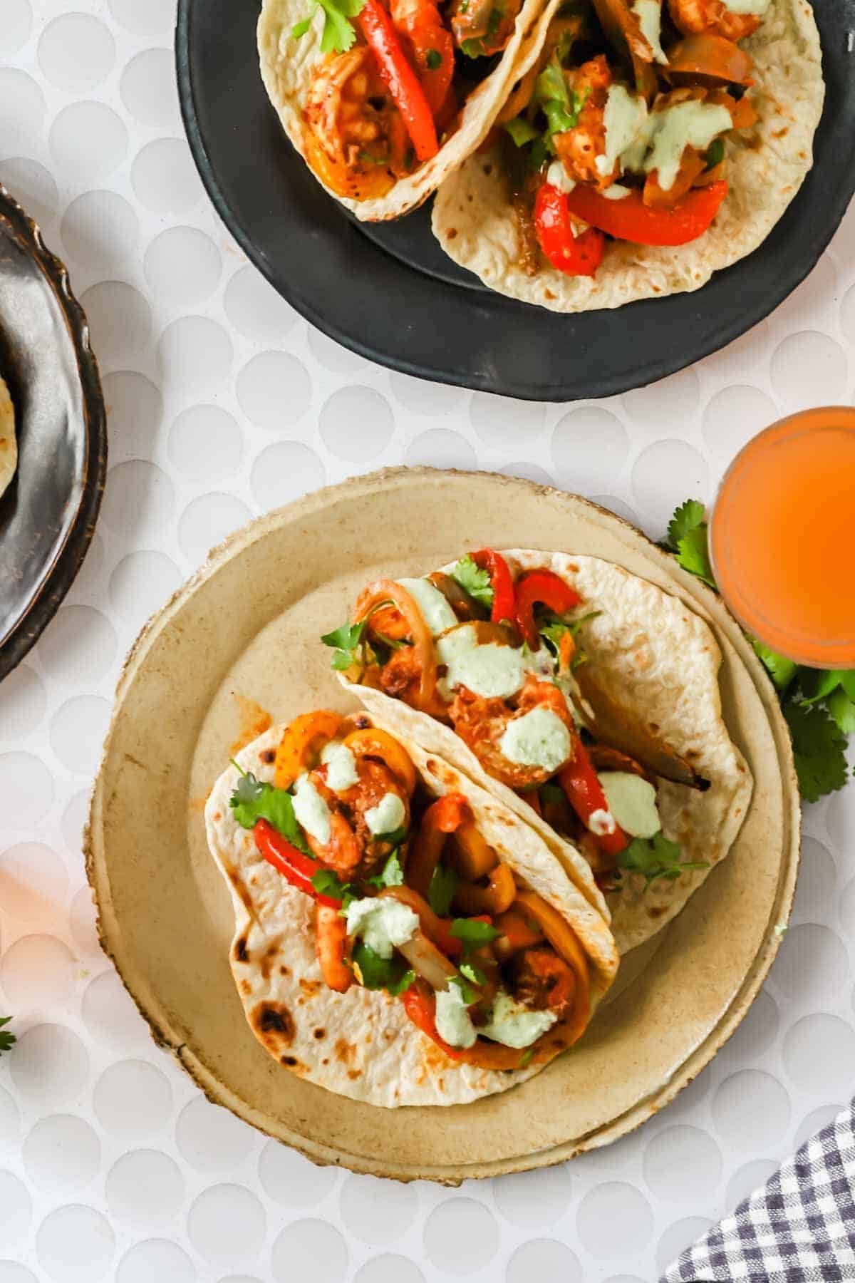 Two flour tortillas filled with shrimp fajitas and drizzled with a creamy sauce are served on a beige plate beside a drink on a white textured surface.
