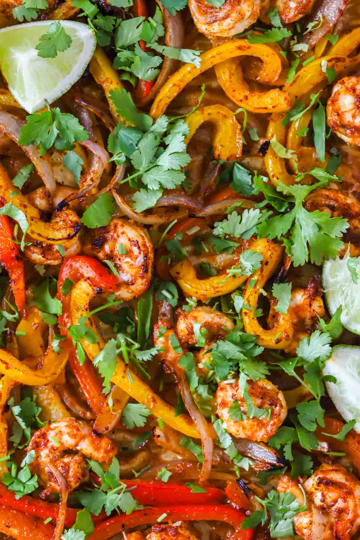 Close-up of cooked shrimp with sliced red and yellow bell peppers, onions, fresh cilantro, and lime wedges.