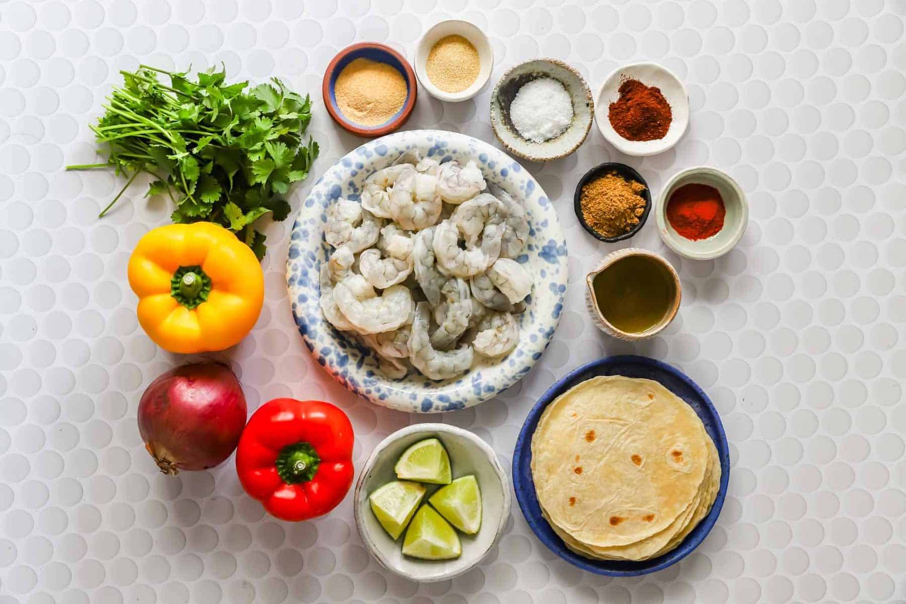 A flat lay of shrimp, tortillas, lime wedges, yellow and red bell peppers, red onion, cilantro, and small bowls of spices and oil on a white textured surface.