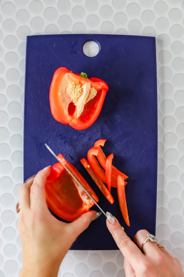 A person slices a red bell pepper into thin strips on a dark blue cutting board with a white textured background.