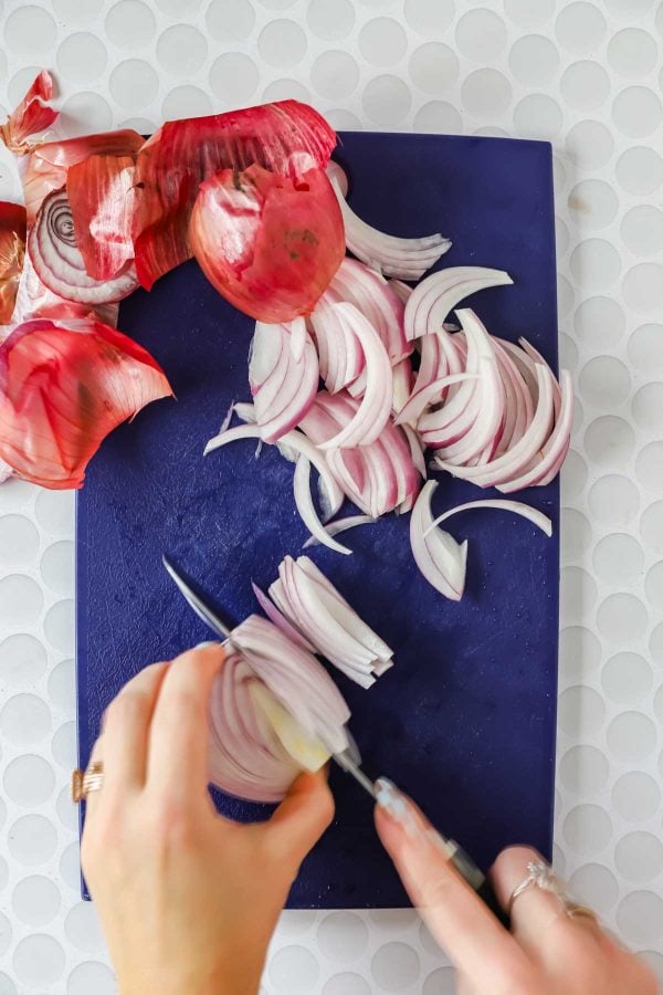 Hands slicing a red onion on a blue cutting board, with onion skins and additional onion slices nearby on a white textured surface.