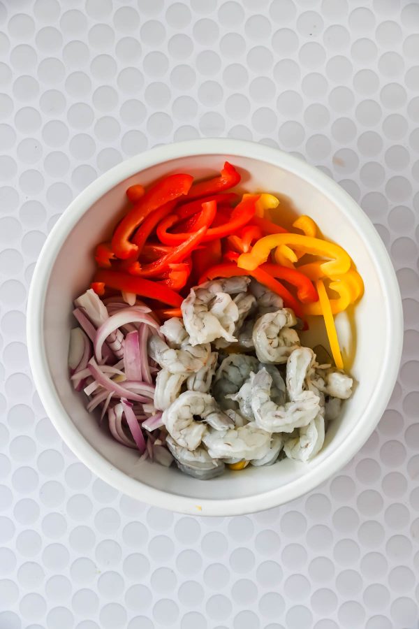 A white bowl containing raw shrimp, sliced red and yellow bell peppers, and sliced red onions on a white textured surface.