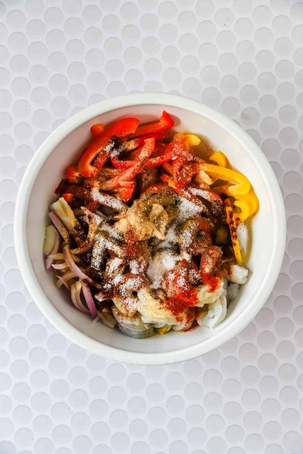 A white bowl filled with sliced red and yellow bell peppers, red onions, and various spices on top, on a white textured surface.
