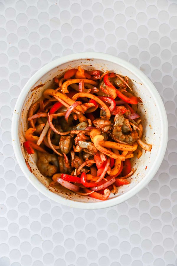 A white bowl filled with sliced red onions, red and yellow bell peppers, and raw shrimp coated in spices, on a white textured surface.