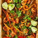 Sheet pan with cooked shrimp, sliced red and yellow bell peppers, onions, lime wedges, and chopped cilantro. Text reads “The Best Sheet Pan Shrimp Fajitas.