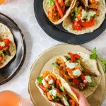 Three shrimp fajitas with red peppers and cilantro are served on tortillas, displayed on plates and a white surface, with a hand holding one and a drink nearby.