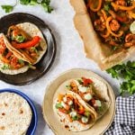 Three plates with tortillas topped with sheet pan shrimp fajitas and sauce, a tray of cooked vegetables, and fresh cilantro on a white surface.