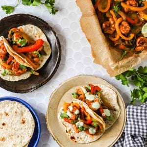 Three plates with tortillas topped with sheet pan shrimp fajitas and sauce, a tray of cooked vegetables, and fresh cilantro on a white surface.