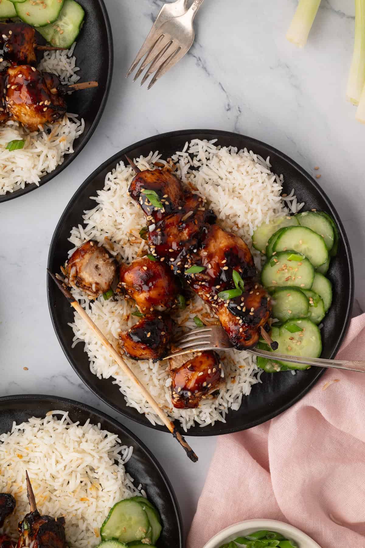 A black plate with air fryer chicken skewers on white rice, garnished with sesame seeds and green onions, with sliced cucumbers on the side.