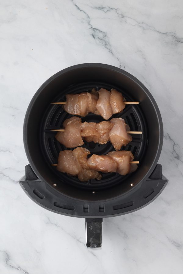 Raw chicken pieces on wooden skewers placed inside an air fryer basket on a marble countertop.