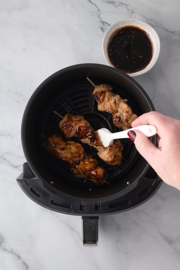 A hand brushes sauce onto chicken skewers in an air fryer basket, with a small bowl of sauce placed nearby on a marble countertop.