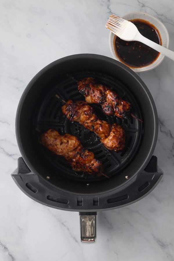 Three pieces of grilled meat on skewers cooking in an air fryer basket, with a bowl of sauce and a brush on the side.