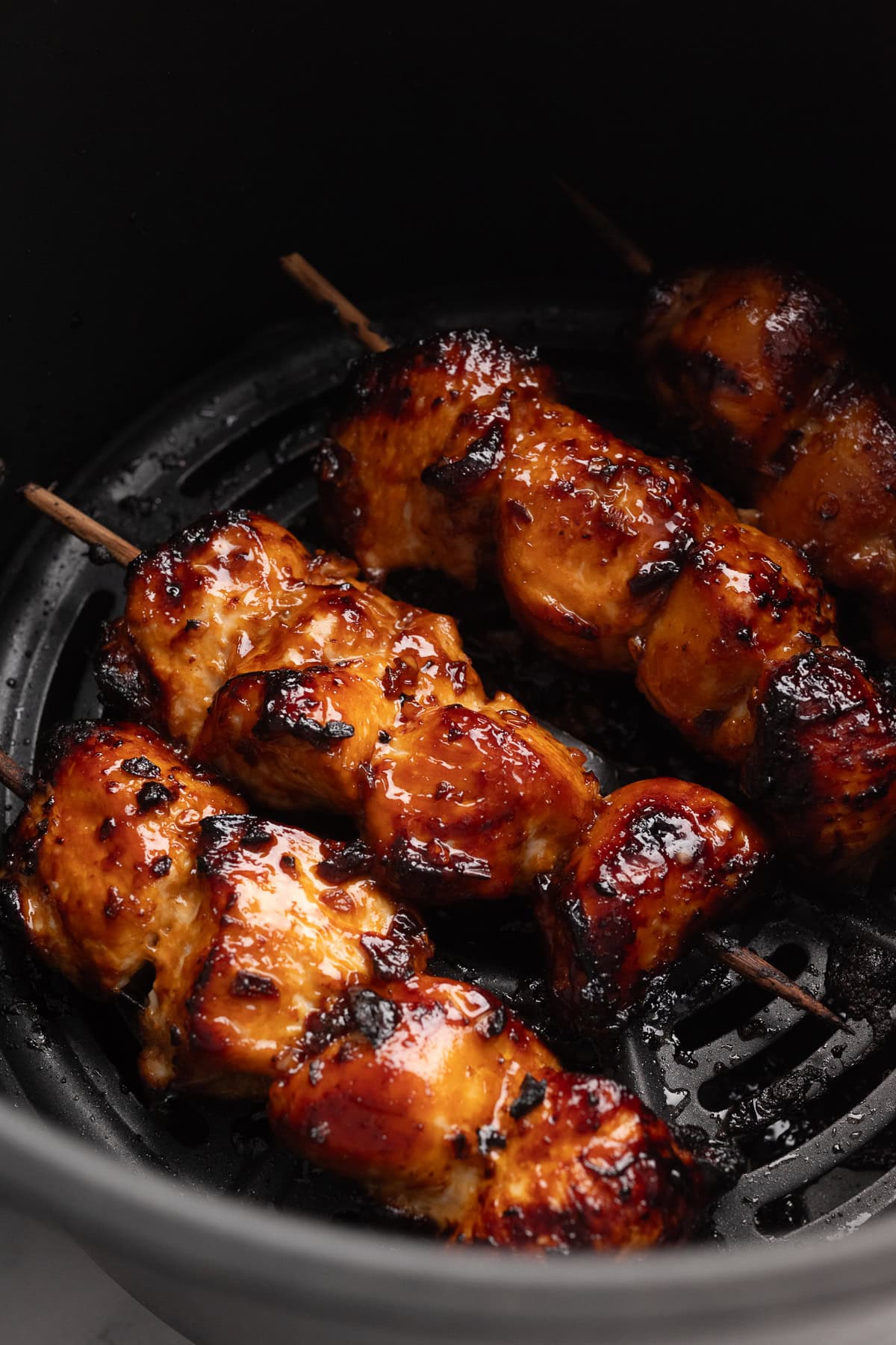 Three skewers of glazed, grilled chicken cooking inside an air fryer basket, showing a caramelized and slightly charred surface.