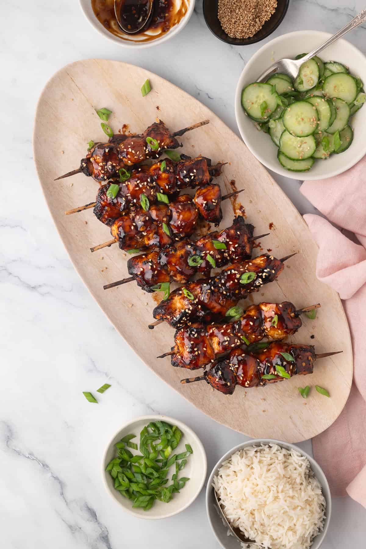 Wooden platter with air fryer chicken skewers glazed in sauce, garnished with green onions; surrounded by bowls of cucumber salad, rice, sesame seeds, and extra green onions on marble surface.