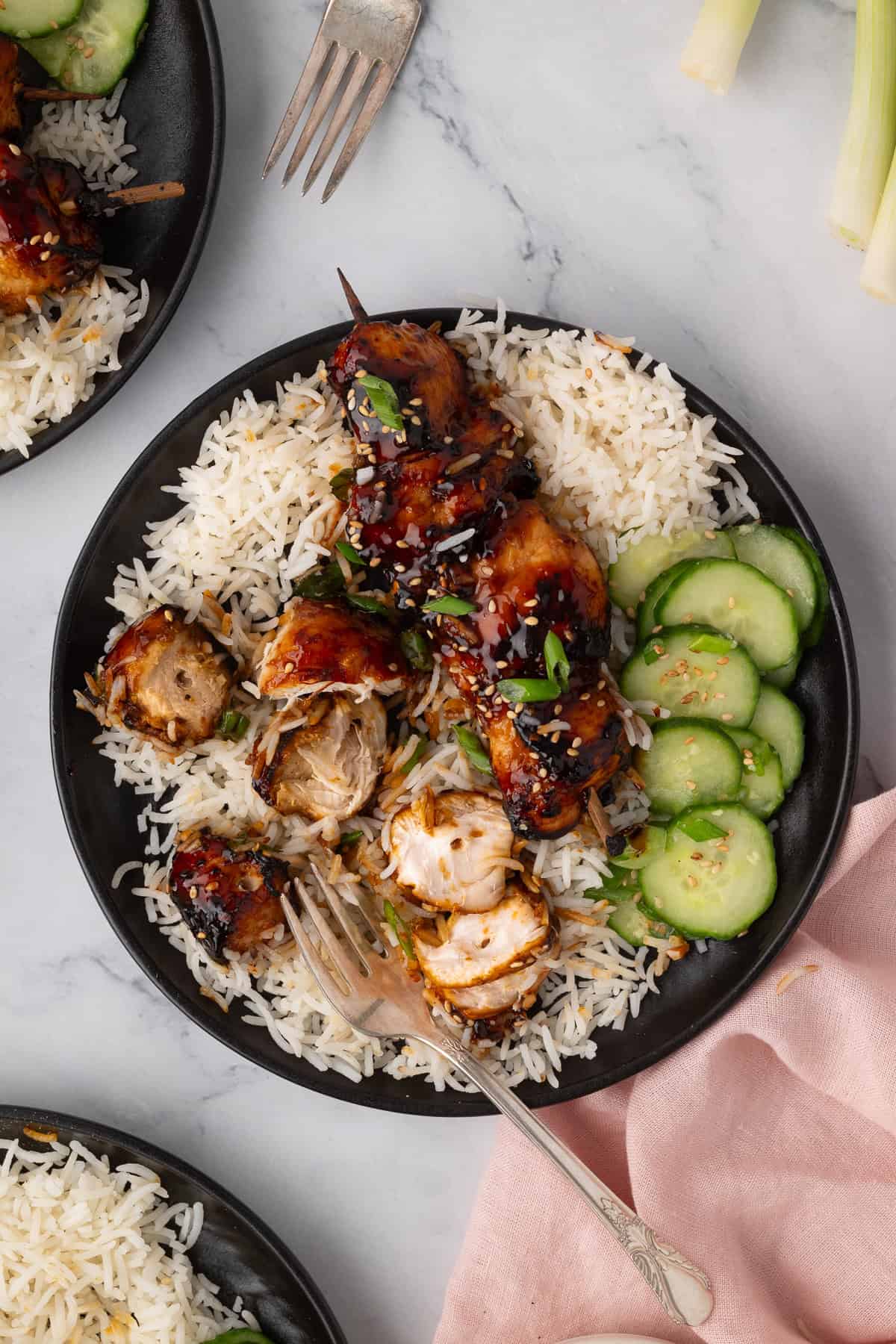 A black plate with grilled chicken skewers on white rice, sliced cucumbers, and a fork, set on a marble surface next to a pink napkin.