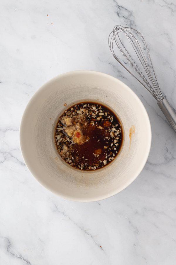 A white bowl containing a mixture of soy sauce, chopped garlic, brown sugar, and other ingredients sits on a marble surface next to a metal whisk.