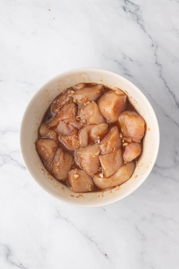 A white bowl filled with raw chicken pieces marinating in a brown sauce sits on a white marble surface.