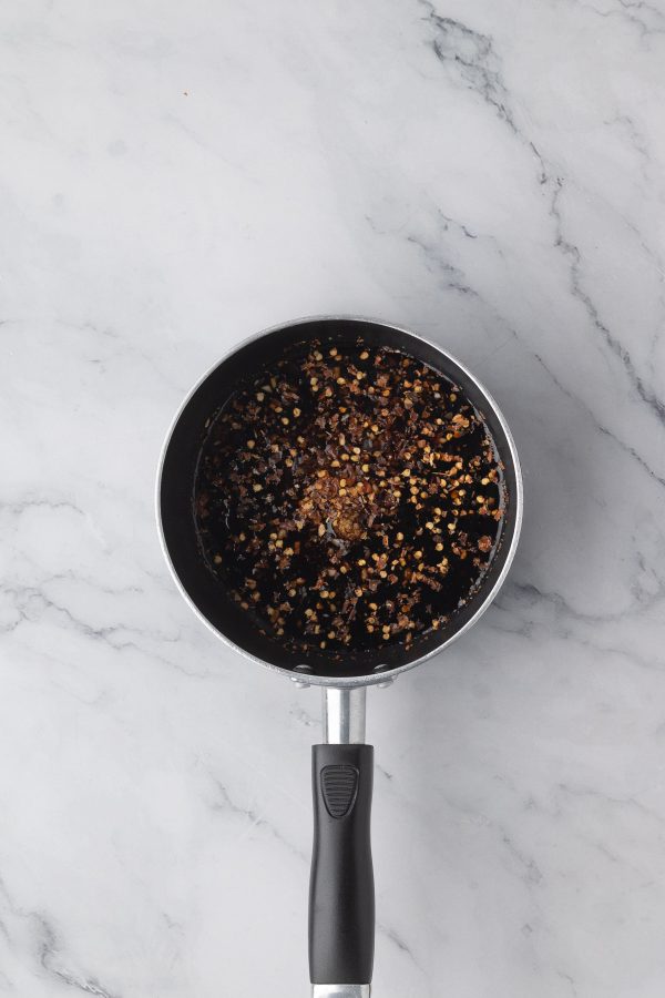 A small saucepan filled with a dark liquid and visible red pepper flakes sits on a light marble surface.