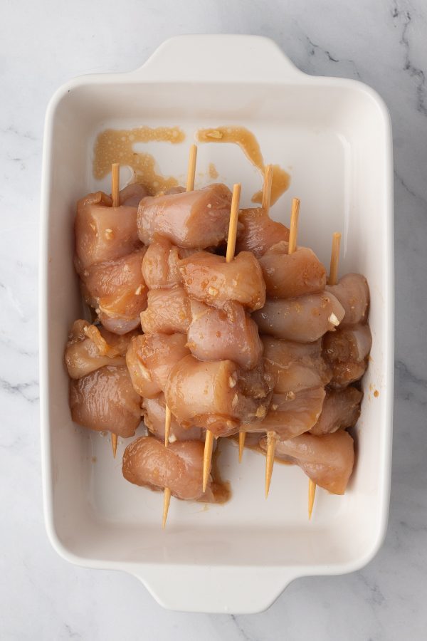 Raw marinated chicken pieces skewered on wooden sticks, arranged in a white rectangular dish on a marble surface.