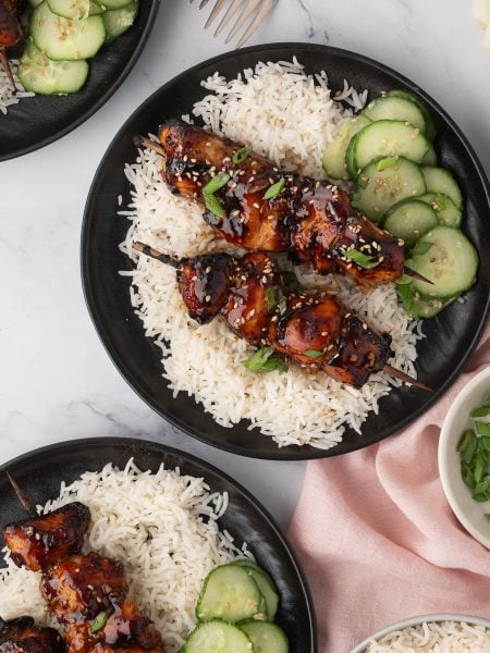 Three plates of rice topped with glazed grilled meat skewers, sliced cucumbers, and garnished with green onions and sesame seeds, arranged on a marble surface.