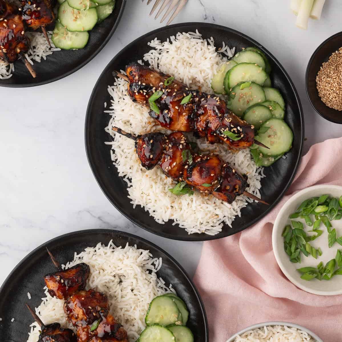 Three plates of rice topped with glazed grilled meat skewers, sliced cucumbers, and garnished with green onions and sesame seeds, arranged on a marble surface.