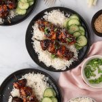 Three black plates with white rice, grilled meat skewers, and sliced cucumbers. Small bowls of sesame seeds and chopped green onions are nearby on a marble surface.