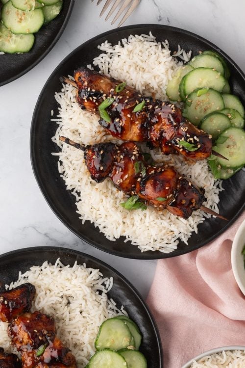 Three black plates with white rice, grilled meat skewers, and sliced cucumbers. Small bowls of sesame seeds and chopped green onions are nearby on a marble surface.