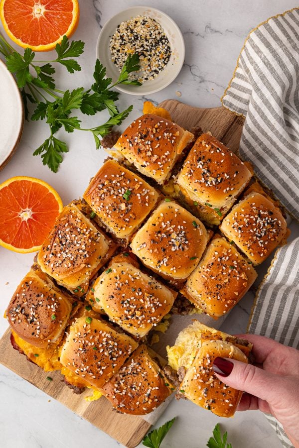 A hand picks up a slider sandwich from a batch of twelve on a wooden board, topped with sesame seeds and surrounded by herbs, orange slices, and a striped towel.