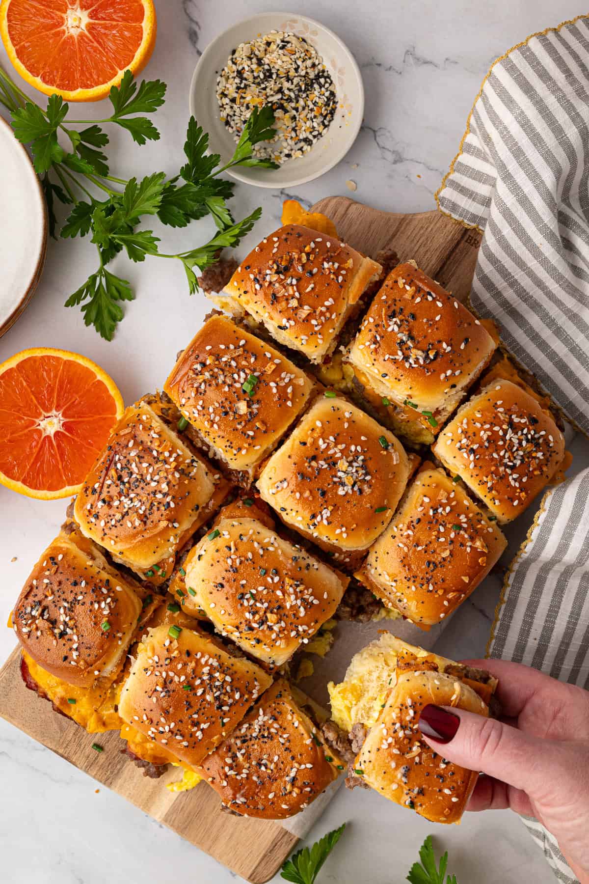A hand picks up a slider sandwich from a batch of twelve on a wooden board, topped with sesame seeds and surrounded by herbs, orange slices, and a striped towel.
