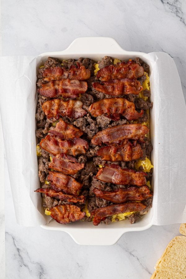 A baking dish lined with parchment paper contains layers of cooked bacon strips and ground sausage over scrambled eggs.
