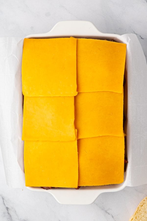 A white baking dish lined with parchment paper contains six slices of cheddar cheese arranged in two rows, covering the contents underneath.