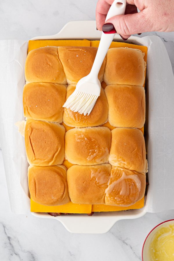 A hand brushes melted butter onto a tray of cheese slider sandwiches in a white baking dish on a marble surface.