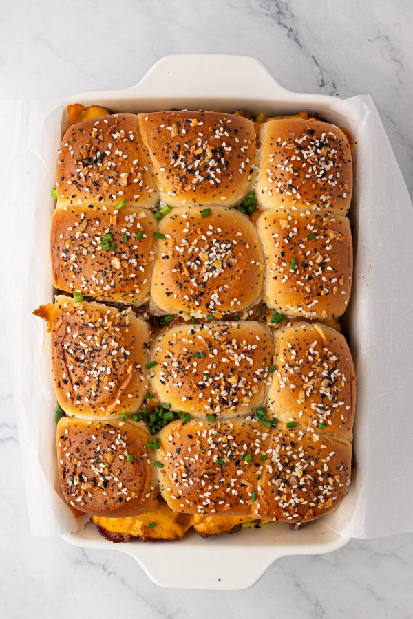 A baking dish containing twelve golden brown slider buns topped with everything bagel seasoning and chopped chives, placed on a marble surface.