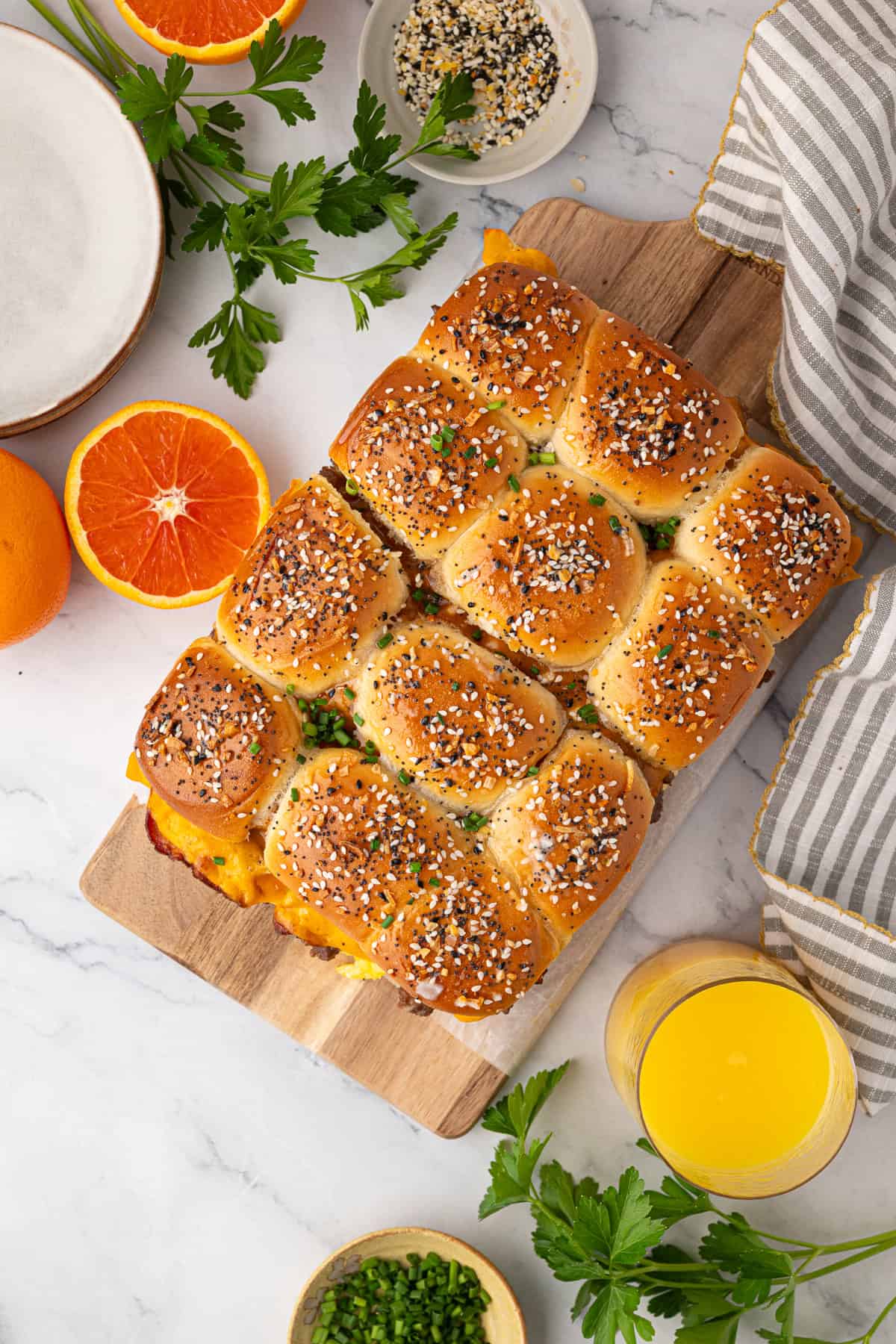 A batch of pull-apart sliders topped with sesame seeds sits on a wooden board, surrounded by fresh herbs, orange slices, and a glass of orange juice.