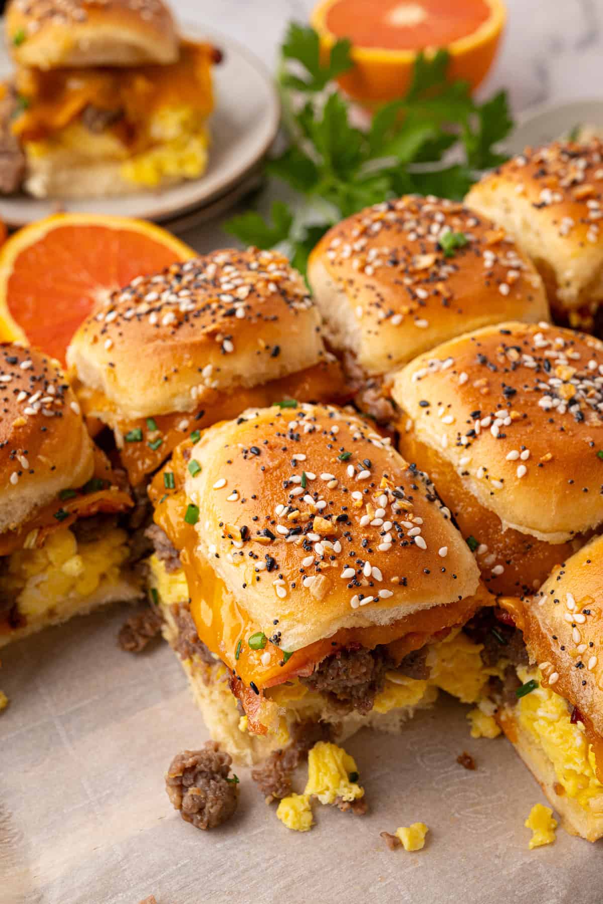 Breakfast sliders made with scrambled eggs, sausage, cheddar cheese, and bacon on everything-seasoned slider buns, arranged on a cutting board with orange slices in the background.
