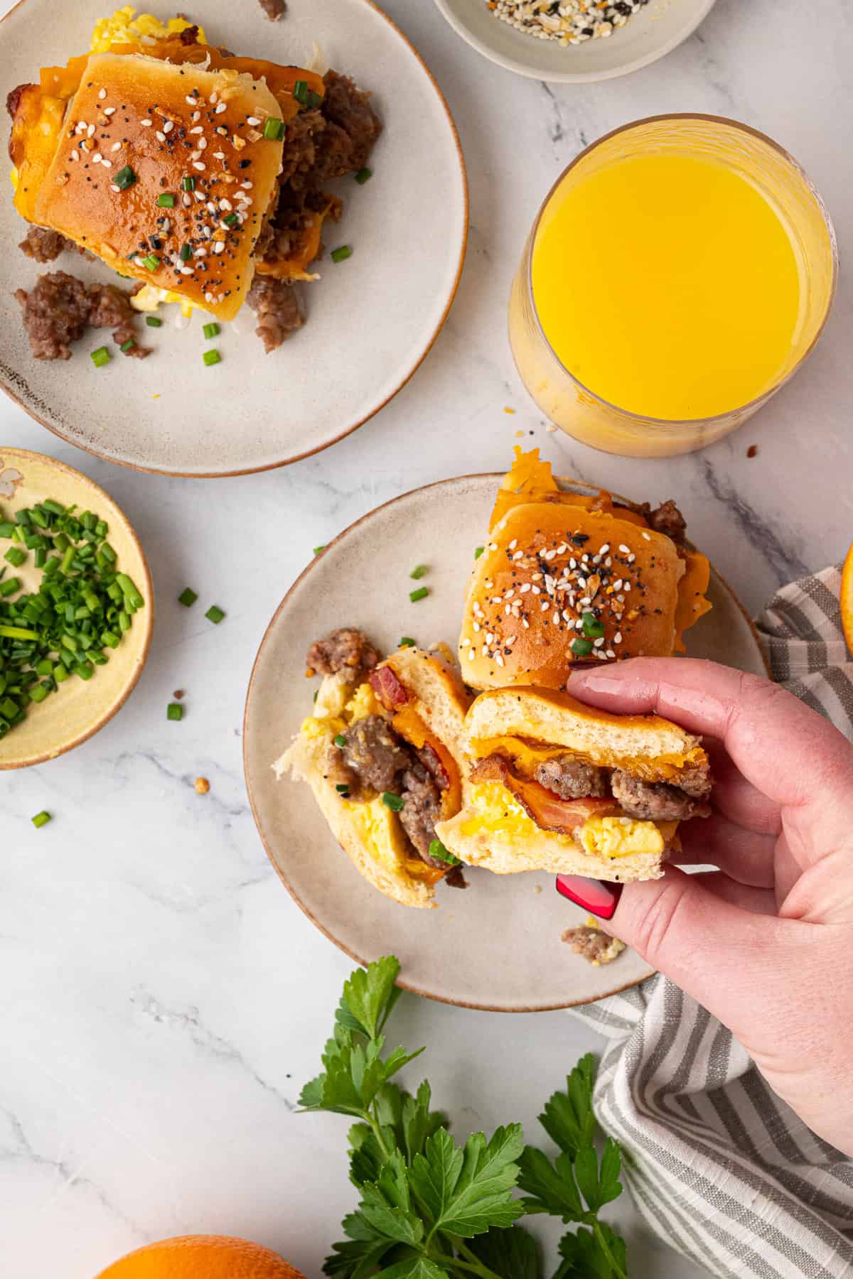 A hand holds a breakfast slider with egg, sausage, and cheese; more sliders, chopped chives, and a glass of orange juice are on a marble table.