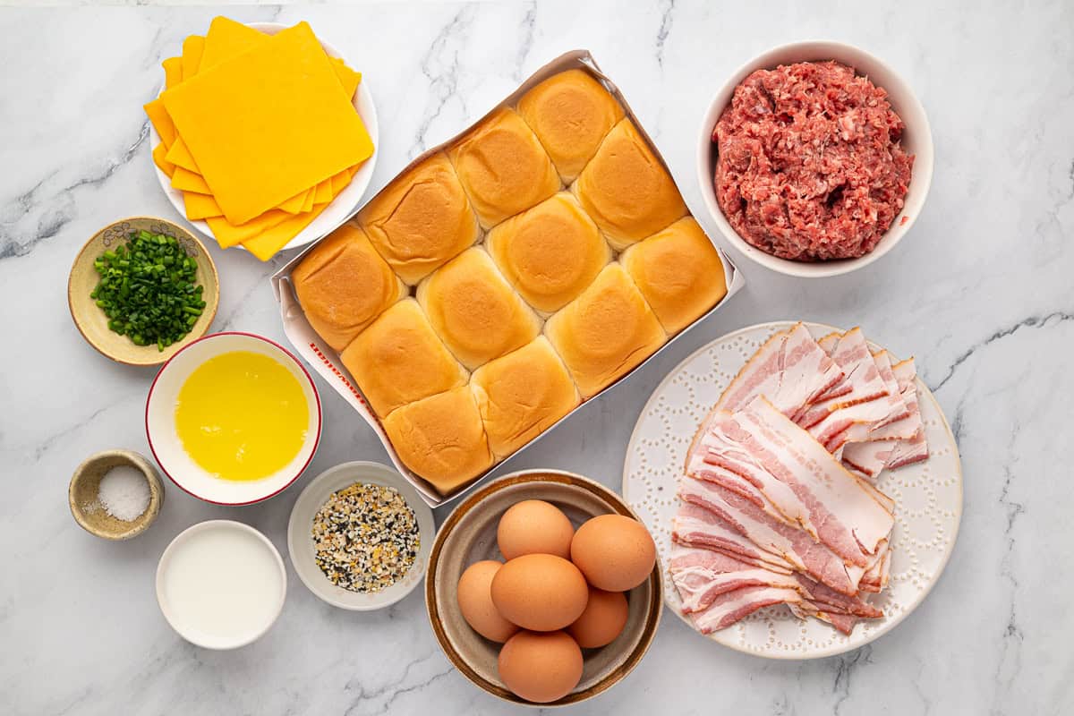 Ingredients on a marble surface including cheddar cheese slices, slider buns, ground beef, bacon, eggs, butter, milk, chopped chives, seasoning, and salt.