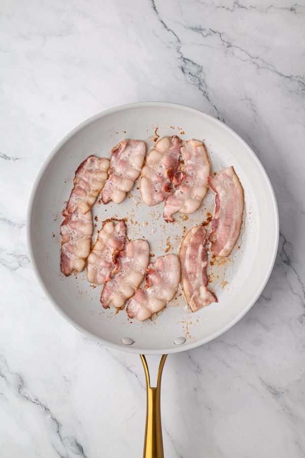 Slices of bacon cooking in a white frying pan with a gold handle on a marble countertop.