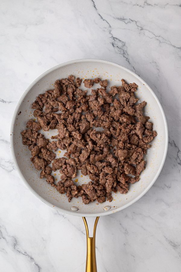 Cooked ground beef in a white frying pan with a gold handle, placed on a light marble countertop.